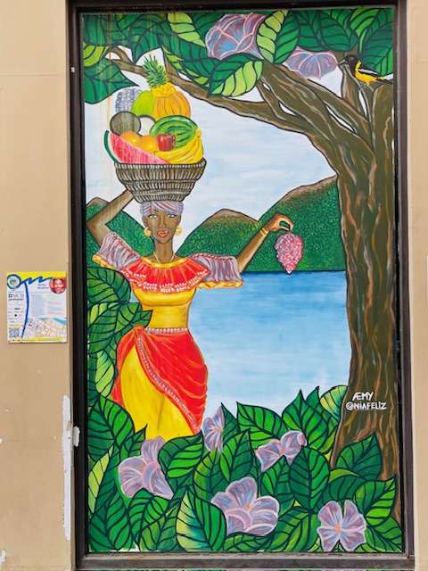 Curacao street art by Amy featuring a native woman in a red, yellow and purple outfit balancing a basket of fruit on her head while walking beside a treet and through a flower garden